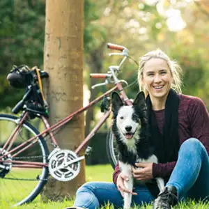 Frau sitzt mit ihrem Hund vor ihrem Fahrrad auf der Wiese und macht eine Pause