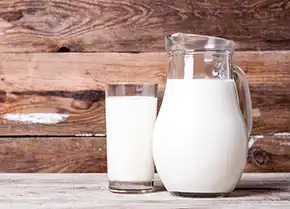 ein gefülltes Glas Milch steht neben einer Milchkanne aus Glas